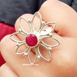 Puede incluir: Un anillo de plata con una piedra preciosa roja en el centro, con forma de flor de seis p&eacute;talos. El anillo est&aacute; en un dedo.
