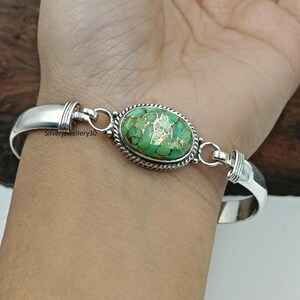Könnte beinhalten: Ein silbernes Armband mit einem ovalen, grün-goldenen Mittelstein. Der Stein ist in einer silbernen Lünette mit einem seilartigen Rand gefasst. Das Armband befindet sich an einem Handgelenk.