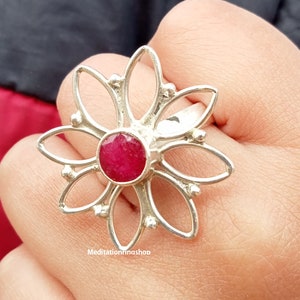 Puede incluir: Un anillo de plata con una piedra preciosa roja en el centro, con forma de flor de seis p&eacute;talos. El anillo est&aacute; en un dedo.