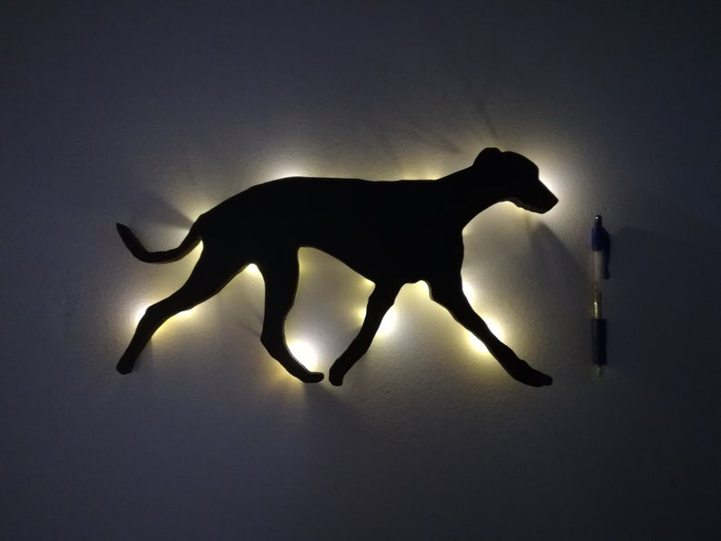May include: A black silhouette of a dog running, lit up with small white lights. The silhouette is mounted on a white wall.