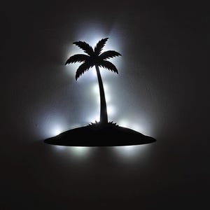 Op de afbeelding: Zwart silhouet van een palmboom op een klein eiland, verlicht door witte lichten. Het eiland en de boom zijn aan een muur bevestigd, wat een tropische sfeer creëert. De verlichting benadrukt de vorm van de boom.