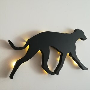 May include: Black silhouette of a dog running, mounted on a white wall with a light shining through the cutout.