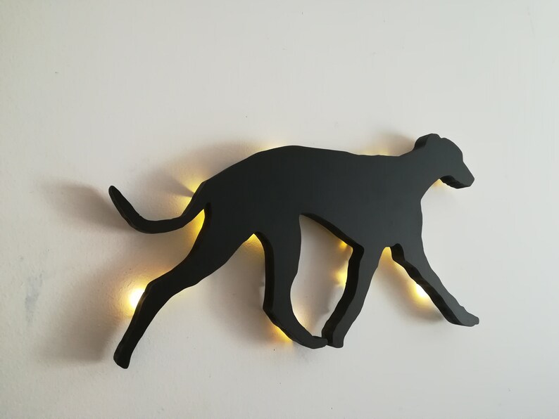 May include: Black silhouette of a dog running, mounted on a white wall with a light shining through the cutout.