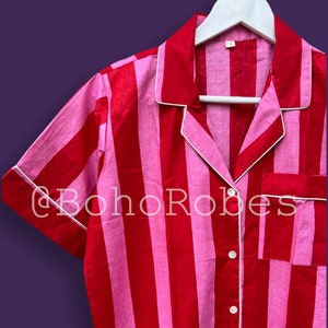Puede incluir: Primer plano de una camisa de pijama a rayas rojas y rosas con ribetes y botones blancos. La camisa está colgada de una percha blanca. El texto "@BohoRobes" es visible en la parte inferior de la imagen.