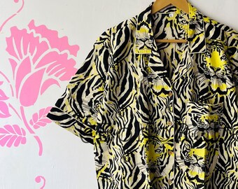 May include: Short-sleeved button-down shirt with a striking tiger print. The shirt features a pattern of black and yellow tiger stripes and tiger faces. The shirt is hanging on a wooden hanger against a white wall with pink floral stencils.