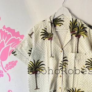 Può includere: Una camicia bianca a maniche corte con bottoni, stampa di palme e pois neri. La camicia ha un colletto e una tasca sul lato sinistro.