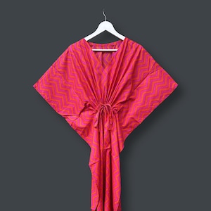 May include: A bright pink and orange patterned kaftan dress with a V-neck and drawstring waist. The dress is hanging on a white hanger against a dark grey background. The fabric features a zigzag design.