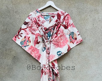 May include: A white cotton kaftan with a pink and green floral print. The kaftan has a V-neck and a tie at the waist. The kaftan is hanging on a white hanger against a gray wall. @boho_robes