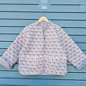 May include: A quilted jacket with a floral pattern in shades of lavender, beige, and gray. The jacket has a stand-up collar, two front pockets, and striped trim on the cuffs and collar. The jacket is hanging on a wooden hanger.