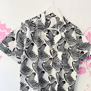 May include: A white button-down shirt with a black and white zebra print. The shirt has short sleeves and a collared neckline.