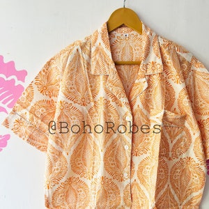 May include: A white short-sleeved button-down shirt with an orange floral pattern. The shirt has a collar and a button-up front. The shirt is made of a lightweight fabric and is perfect for warm weather.