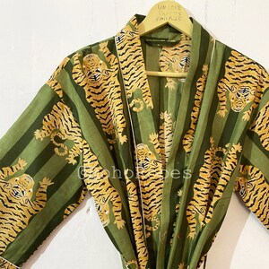 Peut inclure: Robe rayée verte et dorée avec un imprimé tigre. La robe a une ceinture nouée et une bordure en satin. Le texte "UNIQUE CRAFTS FABRIC" est écrit sur un cintre en bois.