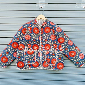 May include: A quilted jacket with a vibrant floral pattern. The jacket features a blue background with large red flowers and green leaves. The edges are trimmed with a striped fabric. The jacket is hanging on a wooden hanger.