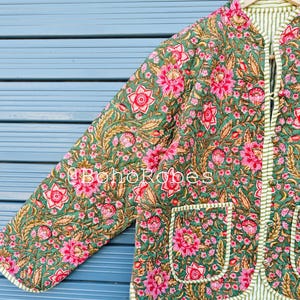 Peut inclure: Veste matelassée à motifs floraux verts, roses et dorés. La veste a une doublure et une bordure rayées, avec deux poches avant. Le texte "@BohoRobes" est visible sur la veste.