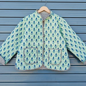 May include: A light green quilted jacket with a blue floral pattern. The jacket has a mandarin collar, two front pockets, and a button-up closure. The cuffs and trim feature a black and white striped pattern.