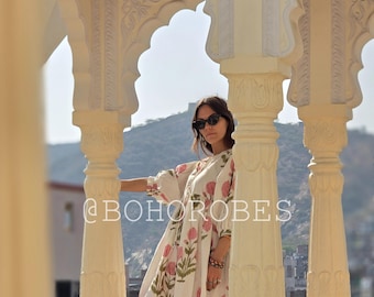 May include: A woman wearing a white dress with a pink floral print stands in a white archway. The dress has a gathered waist and a long, flowing skirt. The woman is wearing sunglasses and has her hair pulled back.