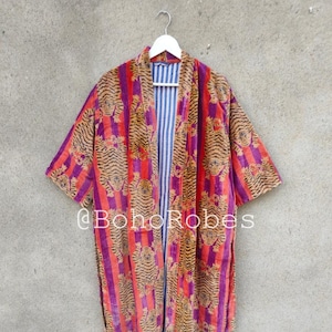 Puede incluir: Una larga bata de rayas moradas y naranjas con estampado de tigre. La bata tiene un cuello ancho y mangas tres cuartos. La bata está colgada de una percha blanca contra una pared gris. El texto "@boho robes" es visible en la bata.