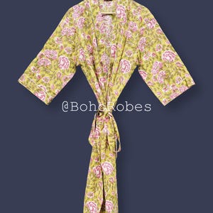 Può includere: Accappatoio con stampa floreale gialla e rosa, appeso a una gruccia di legno. L'accappatoio presenta un motivo ripetuto di fiori rosa su sfondo giallo. Il testo "@BohoRobes" è visibile in basso.