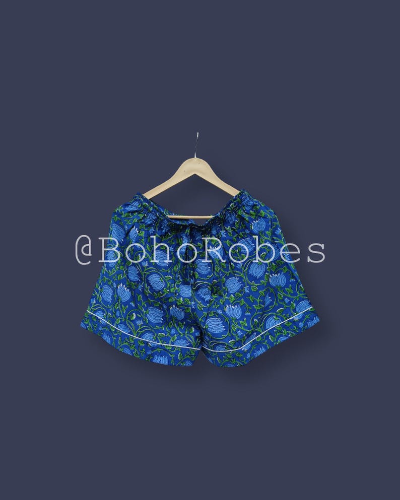 Puede incluir: Shorts azules con estampado floral y ribete blanco, colgados de una percha de madera. Los shorts tienen cintura con cord&oacute;n y un estampado de flores azules y hojas verdes. El texto "@BohoRobes" es visible.