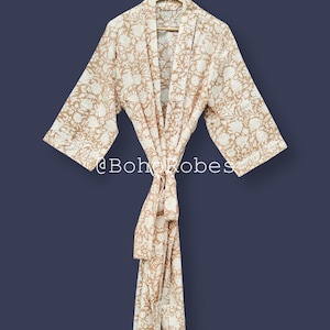 Puede incluir: Bata con estampado floral beige y blanco con cierre de lazo. La bata está colgada de una percha de madera sobre un fondo azul oscuro. El texto "@BohoRobes" es visible en la parte inferior de la bata.