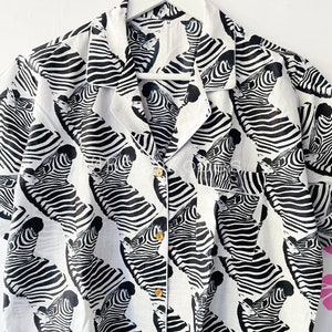 May include: A white button-up shirt with a black and white zebra print. The shirt has short sleeves and a collared neckline.