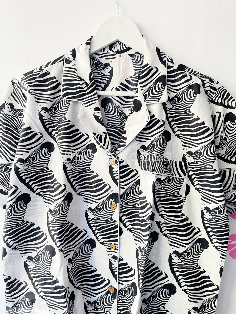 May include: A white button-up shirt with a black and white zebra print. The shirt has short sleeves and a collared neckline.