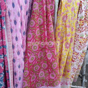 May include: Four colourful cotton scarves with floral patterns. The scarves are pink, red, yellow, and white with different floral designs. The scarves are hanging on a wooden structure.