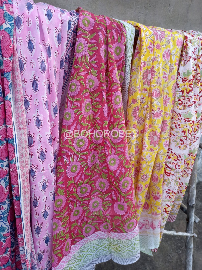 May include: Four colourful cotton scarves with floral patterns. The scarves are pink, red, yellow, and white with different floral designs. The scarves are hanging on a wooden structure.
