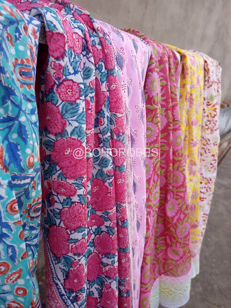 May include: Five colourful fabric scarves with floral patterns. The scarves are hanging on a line and are made of a soft, lightweight material. The scarves are in shades of pink, blue, yellow, and green.