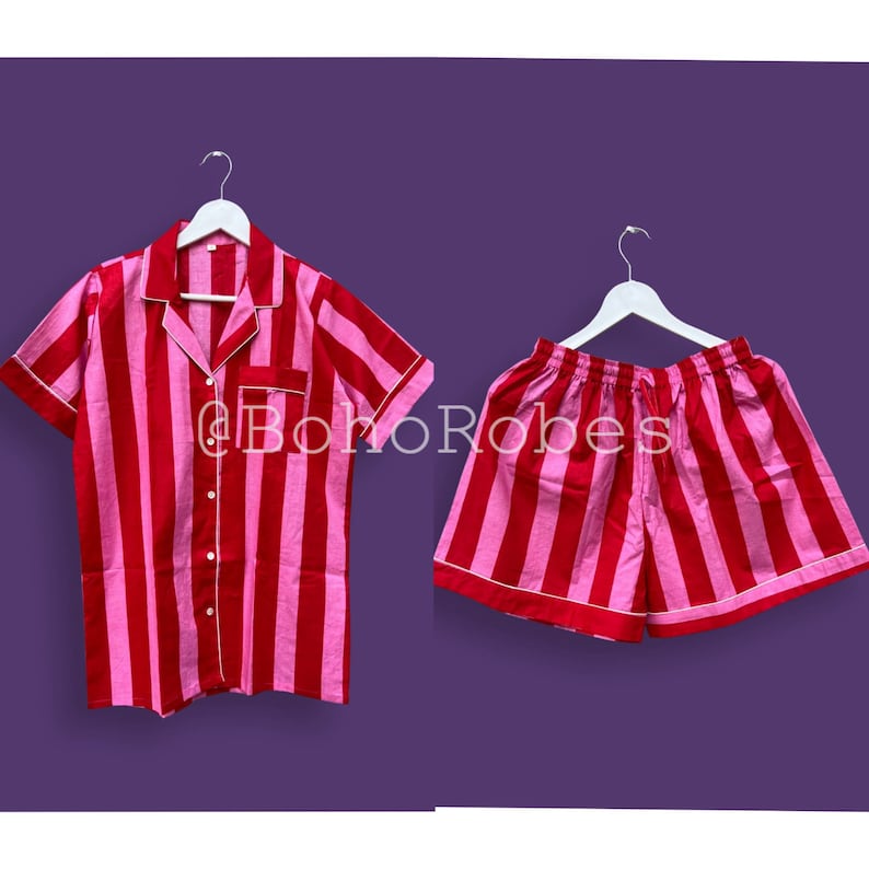 K&ouml;nnte beinhalten: Ein gestreiftes Pyjama-Set in Pink und Rot mit einem kurz&auml;rmeligen Hemd mit Kn&ouml;pfen und einer passenden Hose. Das Set h&auml;ngt an zwei wei&szlig;en Kleiderb&uuml;geln vor einem violetten Hintergrund.
