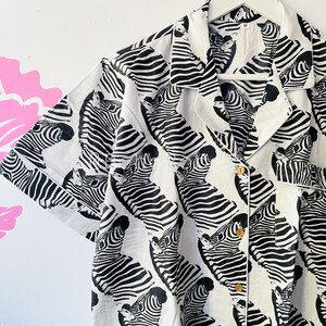 May include: A white button-down shirt with a black and white zebra print. The shirt has a collar and short sleeves.