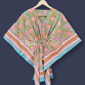 以下が含まれることがあります： 木製のハンガーに掛けられた、ピンクとゴールドの花柄のターコイズ色のカフタン。 カフタンはVネックとドローストリングウエストを備えています。 エッジはストライプのボーダーでトリミングされています。