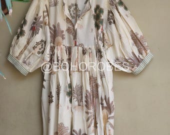 May include: A white cotton dress with a green and brown floral print. The dress has a V-neckline, 3/4 sleeves, and a gathered waist. The dress is hanging on a wooden hanger.