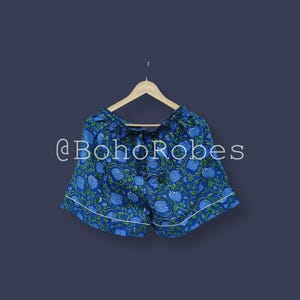 Puede incluir: Shorts azules con estampado floral y ribete blanco, colgados de una percha de madera. Los shorts tienen cintura con cord&oacute;n y un estampado de flores azules y hojas verdes. El texto "@BohoRobes" es visible.