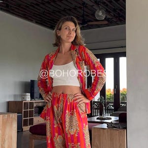 Op de afbeelding: Een vrouw draagt een rode en oranje pyjamaset met een tijgerprint. De set bestaat uit een shirt met lange mouwen en een broek. Het shirt heeft een kraag en een knoopsluiting aan de voorkant. De broek heeft een elastische tailleband. De vrouw staat in een kamer met een houten vloer en een raam op de achtergrond. De tekst "@BOHOROBES" is zichtbaar op het shirt.