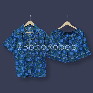 Puede incluir: Conjunto de pijama con estampado floral azul. El conjunto incluye una camisa de manga corta con botones y pantalones cortos a juego, ambos con ribete blanco. La tela presenta un patr&oacute;n repetido de flores azules y hojas verdes. El texto "@BohoRobes" es visible.