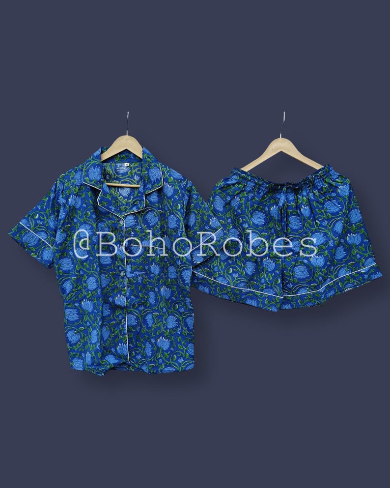 Puede incluir: Conjunto de pijama con estampado floral azul. El conjunto incluye una camisa de manga corta con botones y pantalones cortos a juego, ambos con ribete blanco. La tela presenta un patr&oacute;n repetido de flores azules y hojas verdes. El texto "@BohoRobes" es visible.