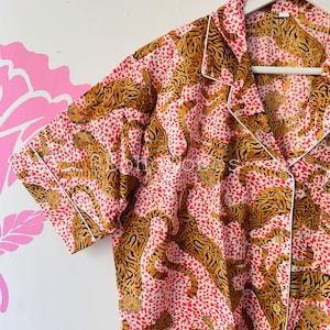 Puede incluir: Una camisa de manga corta rosa y marrón con estampado de leopardo. La camisa tiene un cuello blanco y un cierre de botones en la parte delantera.