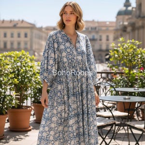 May include: A long, flowing dress with a blue and white floral pattern. The dress has long sleeves and a V-neck. The text "BohoRobes" is printed on the dress. The dress is worn outdoors.