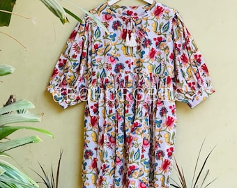 May include: A white cotton dress with a floral print in shades of red, blue, yellow, and green. The dress has a round neckline, 3/4 sleeves, and a tie at the neck with a tassel.