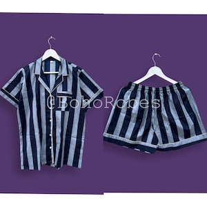 K&ouml;nnte beinhalten: Ein gestreiftes Pyjama-Set mit einem kurz&auml;rmeligen Hemd mit Kn&ouml;pfen und einer passenden Hose. Das Hemd und die Hose sind schwarz und grau mit wei&szlig;en Streifen. Das Set h&auml;ngt an zwei wei&szlig;en Kleiderb&uuml;geln.