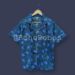Puede incluir: Top de pijama con estampado floral azul con ribete blanco y mangas cortas. La parte superior tiene una parte delantera abotonada y un cuello. El texto "@BohoRobes" est&aacute; impreso en la parte delantera. La camisa est&aacute; colgada en una percha de madera.