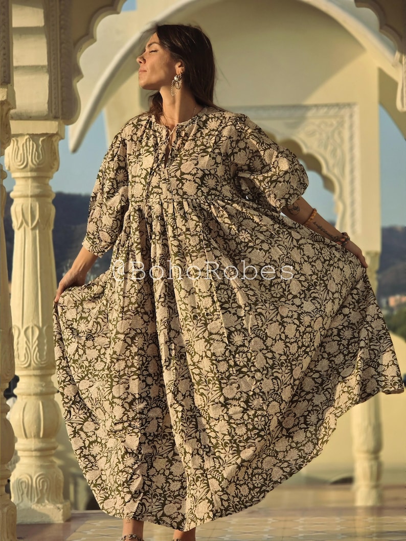 May include: A long, flowing dress featuring a floral pattern in olive green and cream. The dress has a relaxed fit, three-quarter sleeves, and a gathered waist. The text "BohoRobes" is printed on the dress.