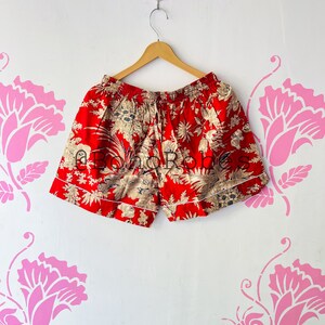 以下が含まれることがあります： 白のトリムが付いた赤とクリーム色の花柄のショートパンツ。ショートパンツは白い壁に木製のハンガーで吊るされています。