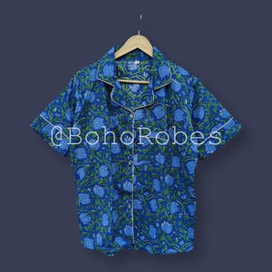 Puede incluir: Top de pijama con estampado floral azul con ribete blanco y mangas cortas. La parte superior tiene una parte delantera abotonada y un cuello. El texto "@BohoRobes" est&aacute; impreso en la parte delantera. La camisa est&aacute; colgada en una percha de madera.