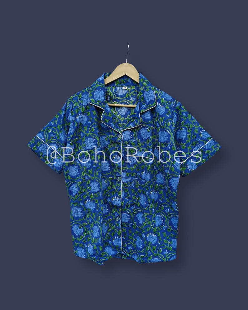 Puede incluir: Top de pijama con estampado floral azul con ribete blanco y mangas cortas. La parte superior tiene una parte delantera abotonada y un cuello. El texto "@BohoRobes" est&aacute; impreso en la parte delantera. La camisa est&aacute; colgada en una percha de madera.