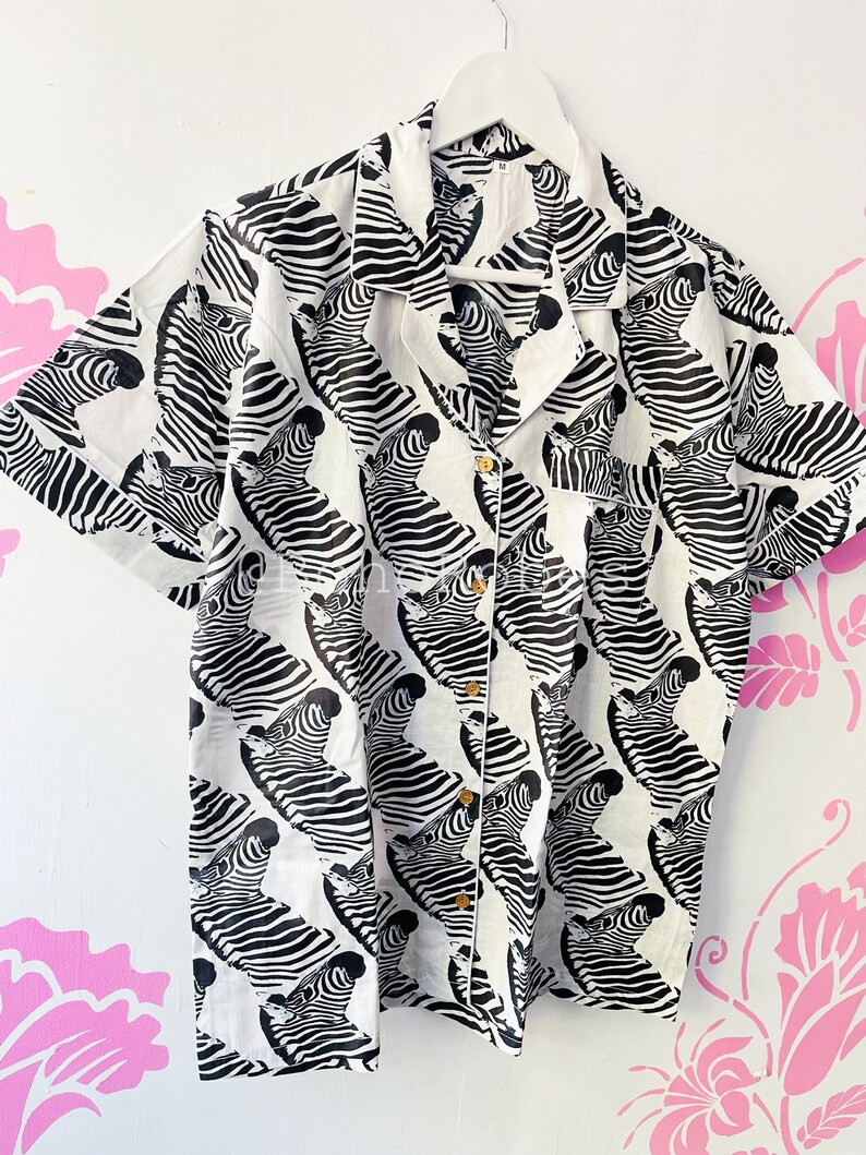 May include: A white button-down shirt with a black and white zebra print. The shirt has short sleeves and a collared neckline.