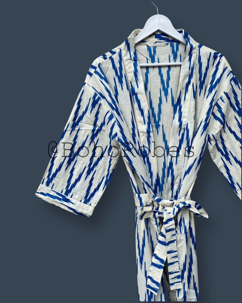 May include: A white cotton robe with a blue ikat pattern. The robe has a tie belt and long sleeves. The robe is hanging on a white hanger.