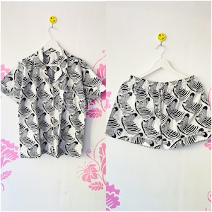 May include: A white and black zebra print short-sleeved button-up shirt and matching shorts. The shirt has a collar and a button-up front. The shorts have an elastic waistband and a drawstring.