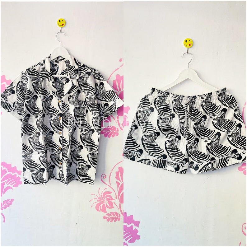 May include: A white and black zebra print short-sleeved button-up shirt and matching shorts. The shirt has a collar and a button-up front. The shorts have an elastic waistband and a drawstring.